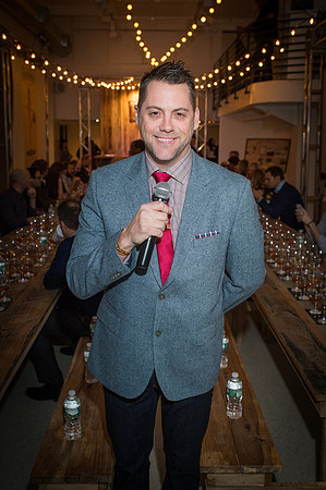Master of Ceremonies holding microphone with tables and string lights behind him.
