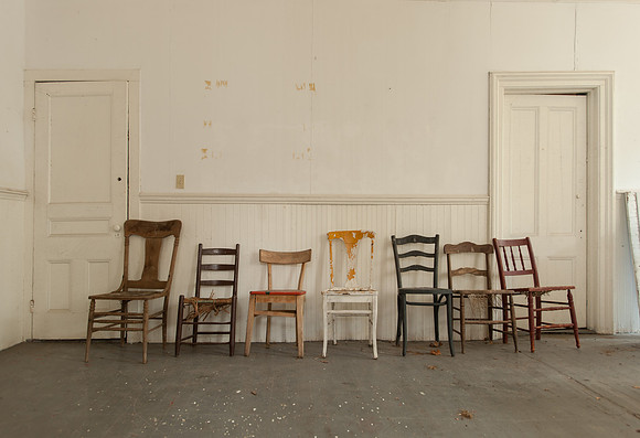 seven different old wooden chairs lined up between two doors against a white wall
