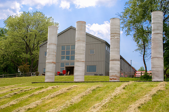 Four freestanding pillars on hill with home and trees behind them. Blue sky.